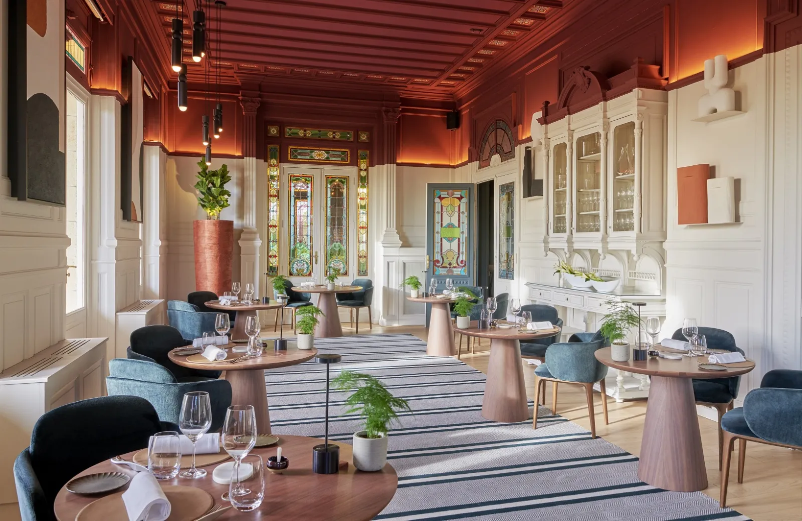 Elegant interior of Grand Barrail restaurant in Bordeaux featuring red wood ceiling, round wooden tables, blue and black chairs, fine glassware, minimalist floral decor, and colorful stained glass windows in background.