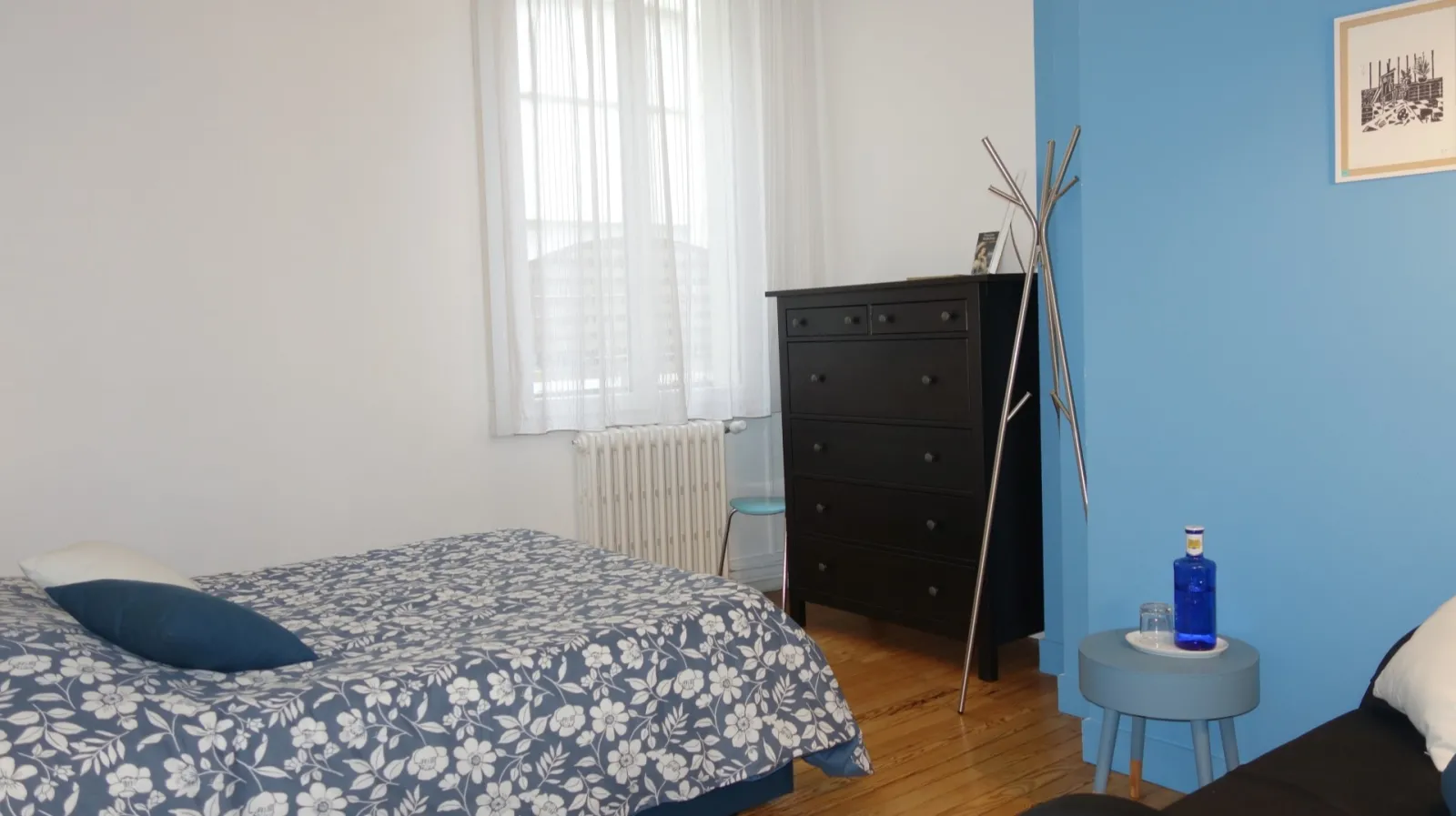 Modern bedroom with blue and white walls, bed with floral patterned cover, black dresser, small grey side table with blue bottle, white radiator and window with white curtains.