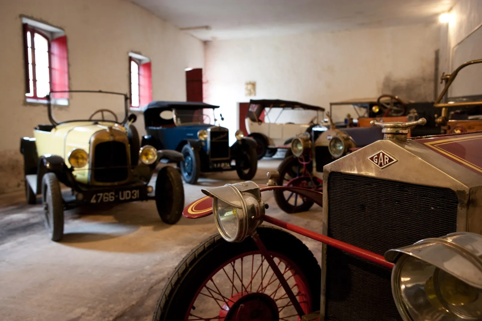 Collection of vintage automobiles and early motorcycles on display in a well-lit museum hangar, featuring classic cars from the early 20th century at Château Carbonnieux.