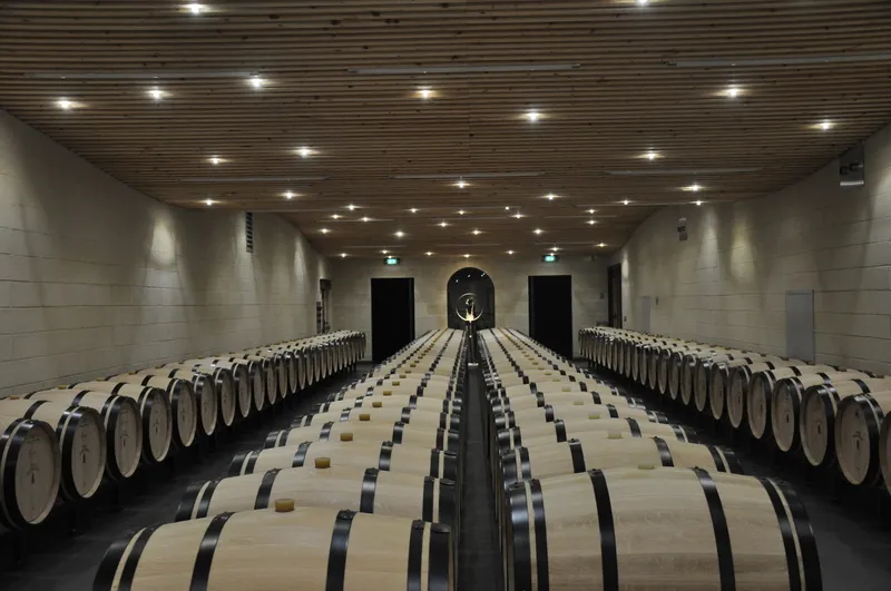 Interior of a wine cellar featuring hundreds of wooden barrels arranged symmetrically along a central corridor, modern ceiling lighting, Château Balestard La Tonnelle in Bordeaux.