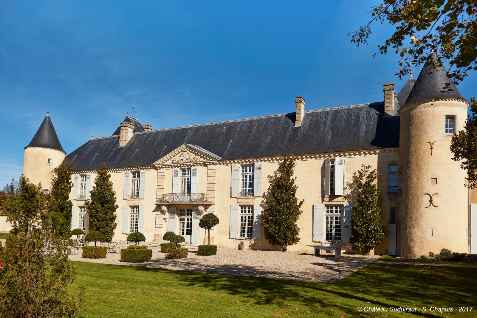 Main façade of Château Suduiraut in Bordeaux, a noble estate with cream-colored walls, round towers with pointed roofs, blue shuttered windows, surrounded by manicured gardens and topiary