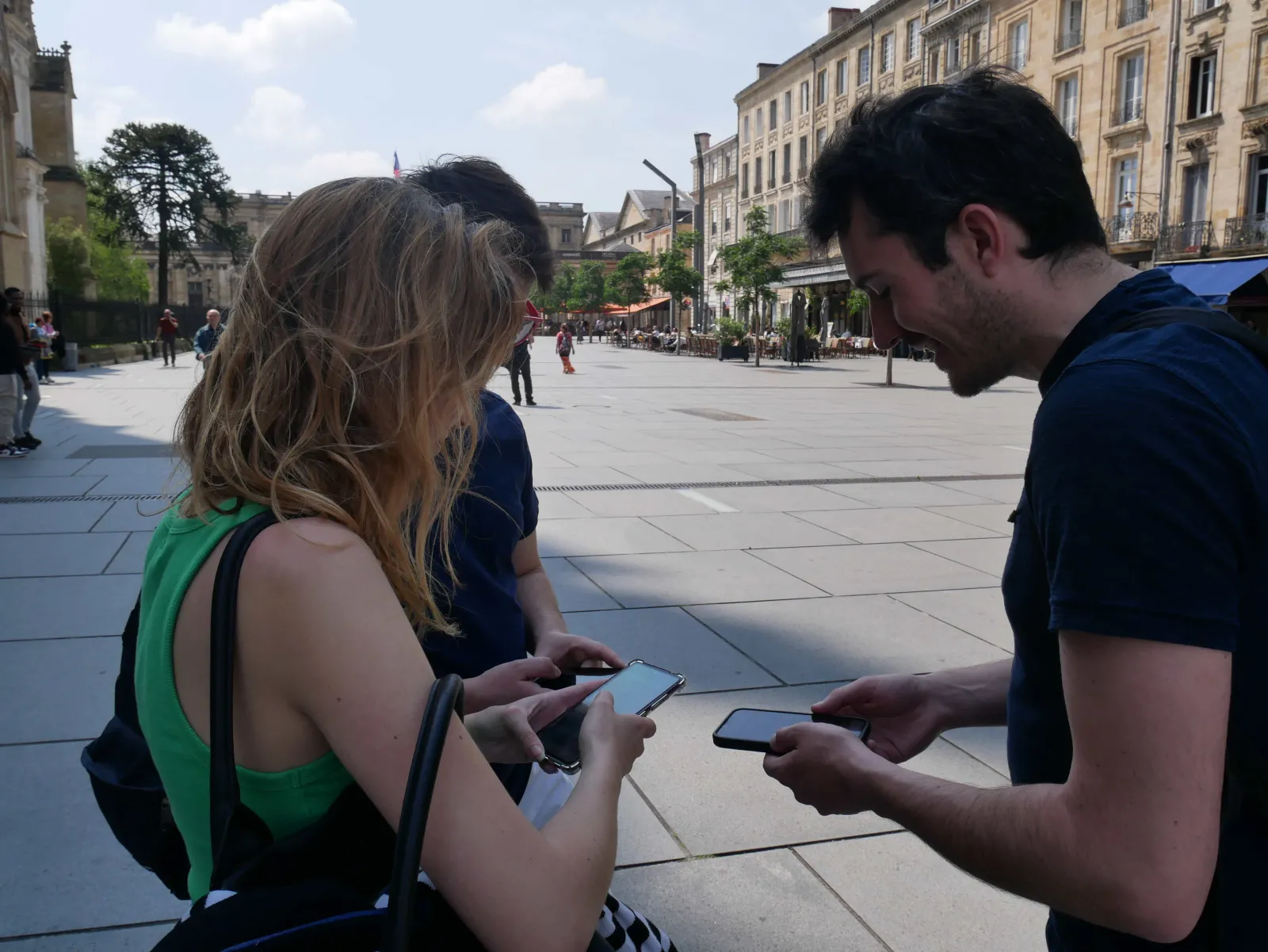 Three people checking their mobile phones on a sunny public square in Bordeaux, with Haussmann-style buildings in the background