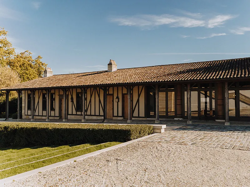 Façade of Château Marquis d'Alesme Becker in Bordeaux, half-timbered house with tiled roof, surrounded by hedges and gravel driveway.