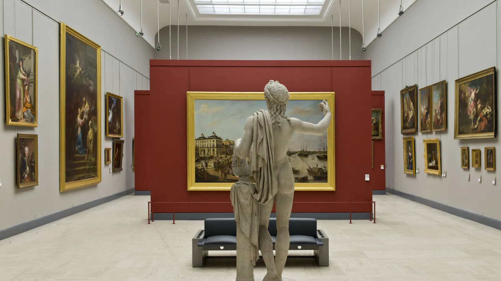 Gallery of the Bordeaux Museum of Fine Arts featuring a marble statue in the foreground facing gilt-framed historical paintings on white and burgundy walls, with overhead lighting.