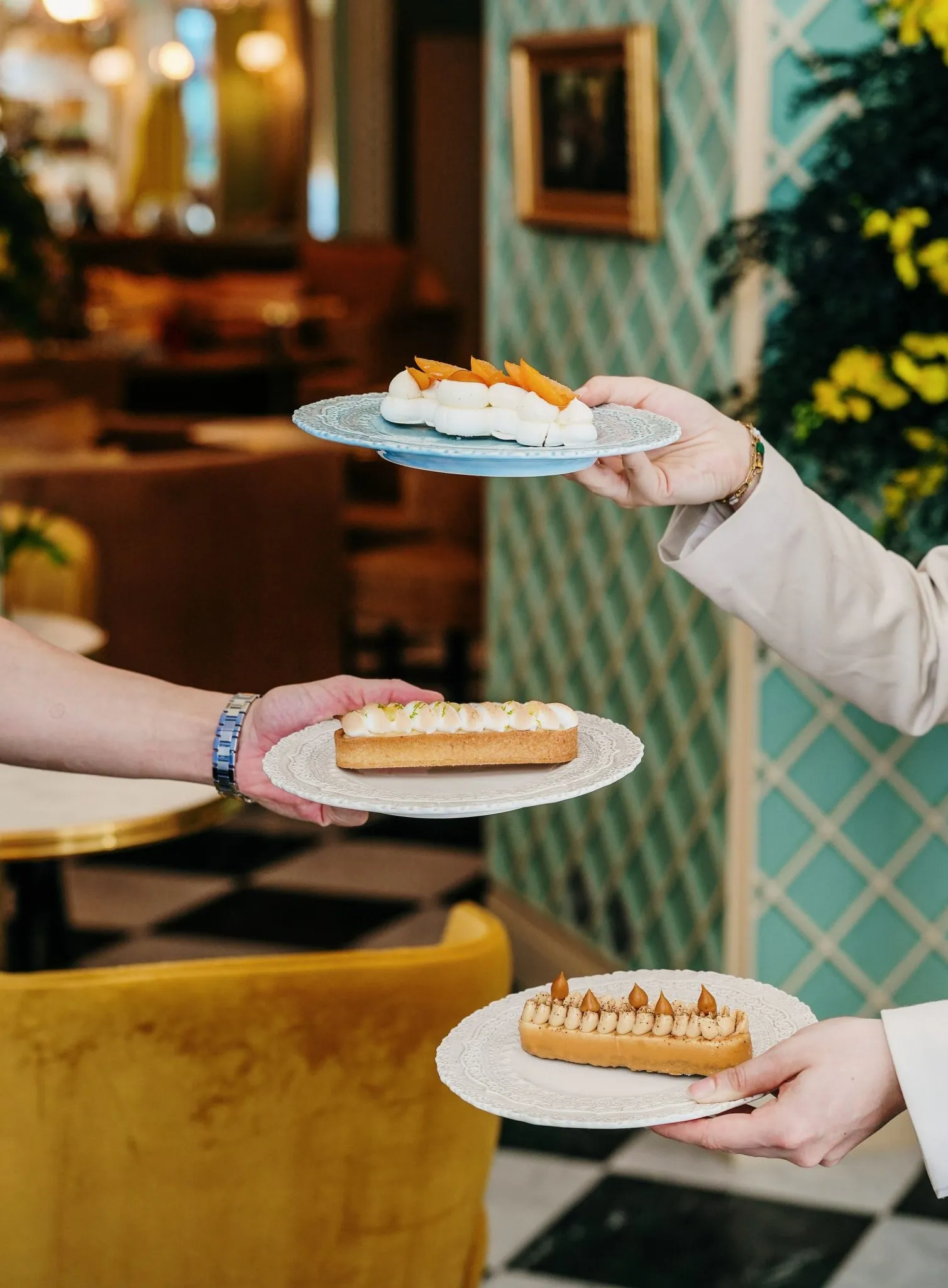 Three hands presenting pastries on white plates in an elegant Bordeaux salon at Le Boudoir de Léa, with turquoise décor and green plants in the background