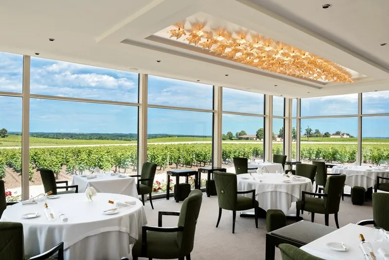 Elegant fine dining restaurant room with round tables set with white linens, black chairs, and floor-to-ceiling windows overlooking Bordeaux vineyards under blue sky.