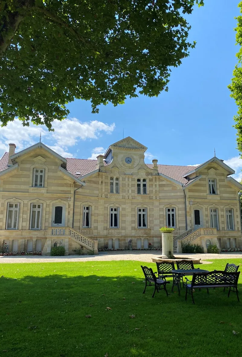 Front view of Château Maucaillou in Bordeaux, a grand pale yellow stone mansion with red tile roof, symmetrical pavilions, set on manicured lawn with garden furniture in the foreground.