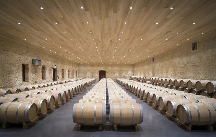 Wine aging cellar at Château Fourcas Hosten in Bordeaux, featuring hundreds of wooden barrels arranged symmetrically beneath a light wood ceiling with integrated lighting