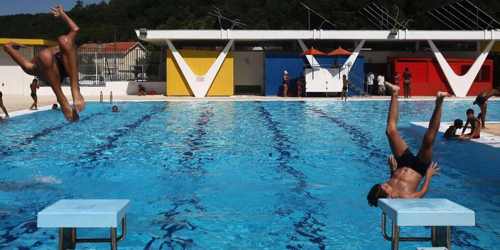 Municipal swimming pool in Floirac, Bordeaux, with multiple swimmers and divers enjoying the turquoise water, colorful buildings visible in the background
