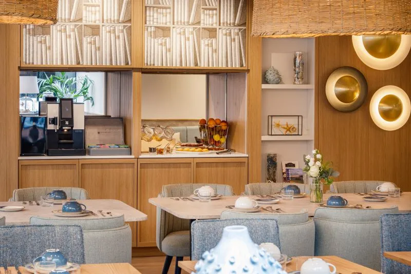 Modern restaurant dining area featuring wooden tables, gray upholstered chairs, light wood wall paneling, circular brass pendant lights, integrated shelving with white books, and open kitchen in the background.