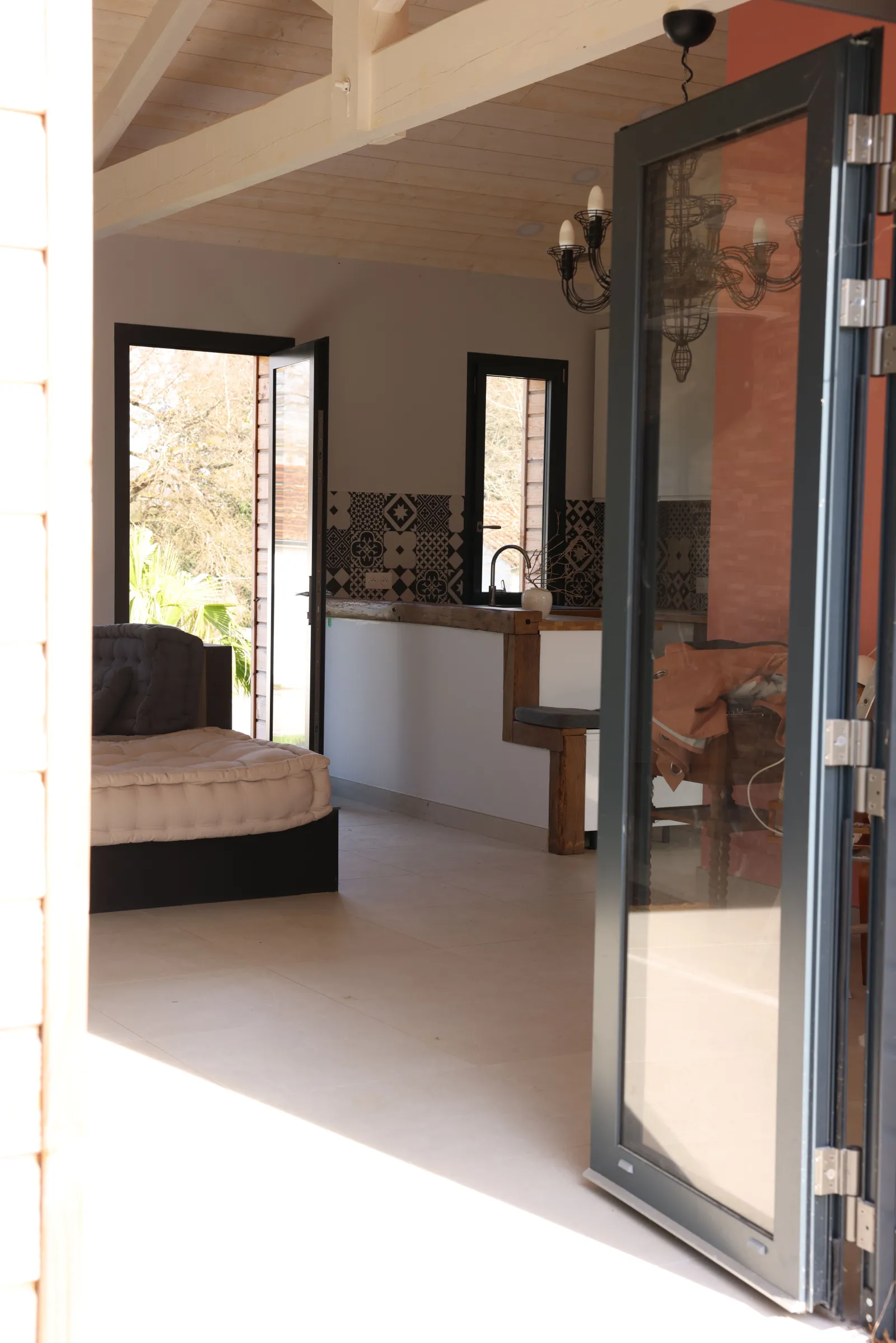 Modern interior of an eco-cottage featuring an open bathroom with wooden sink, framed mirror, sliding glass doors opening onto a garden, and living space with bed visible in the background in Bordeaux.