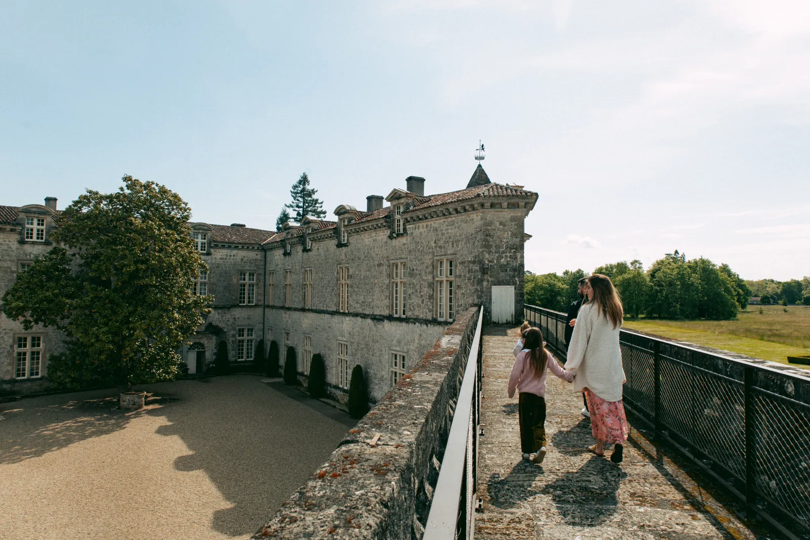 A woman and child walking on the ramparts of Château Royal de Cazeneuve, with the stone castle facade in the background and green fields stretching beyond.