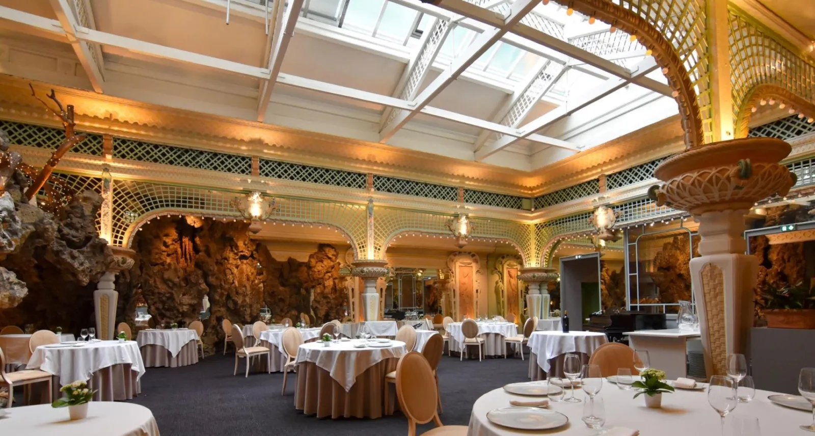 Elegant dining room at Chapon Fin in Bordeaux featuring round tables set with white linens, golden chairs, ornate columns, glass ceiling and geometric gold-patterned wall decorations