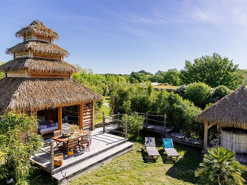Aerial view of an eco-tourism resort featuring thatched-roof bungalows, wooden decks, pool, and lush green surroundings near Bordeaux