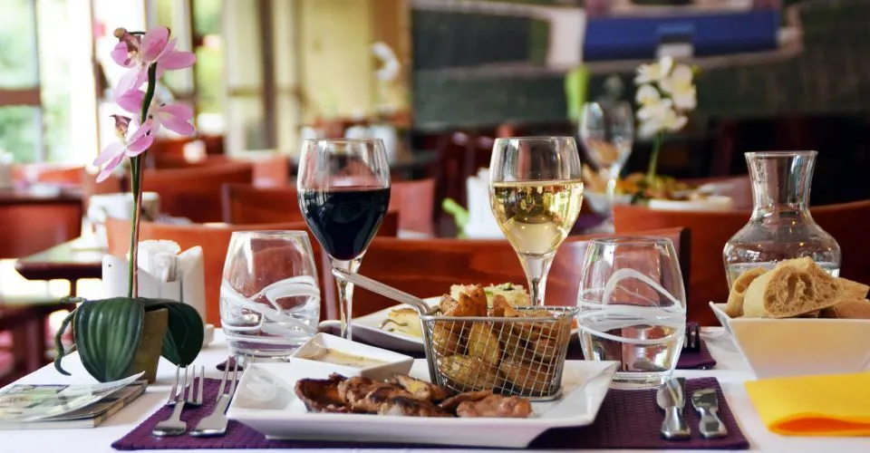 Elegant dining table set with red and white wine glasses, charcuterie plate, toasted bread basket, pink flower in vase, and cutlery on purple tablecloth in an upscale restaurant dining room.