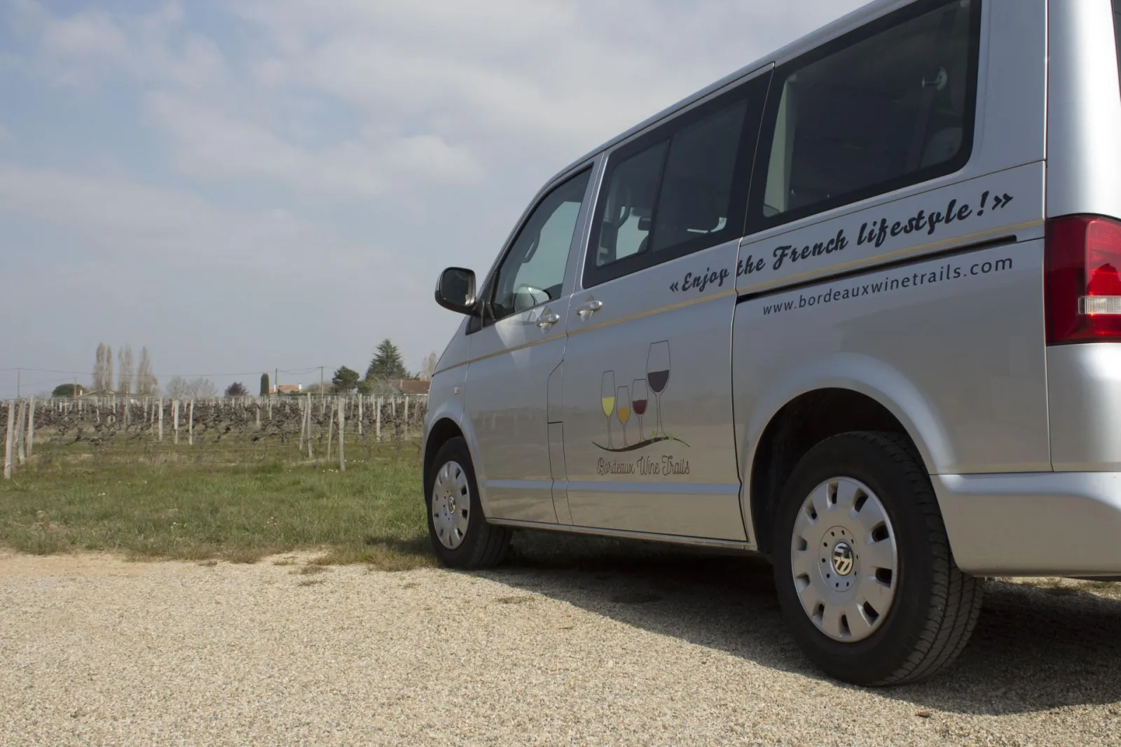 A grey Bordeaux Wine Trails minibus parked in a Bordeaux vineyard with rows of vines and support posts visible in the background.