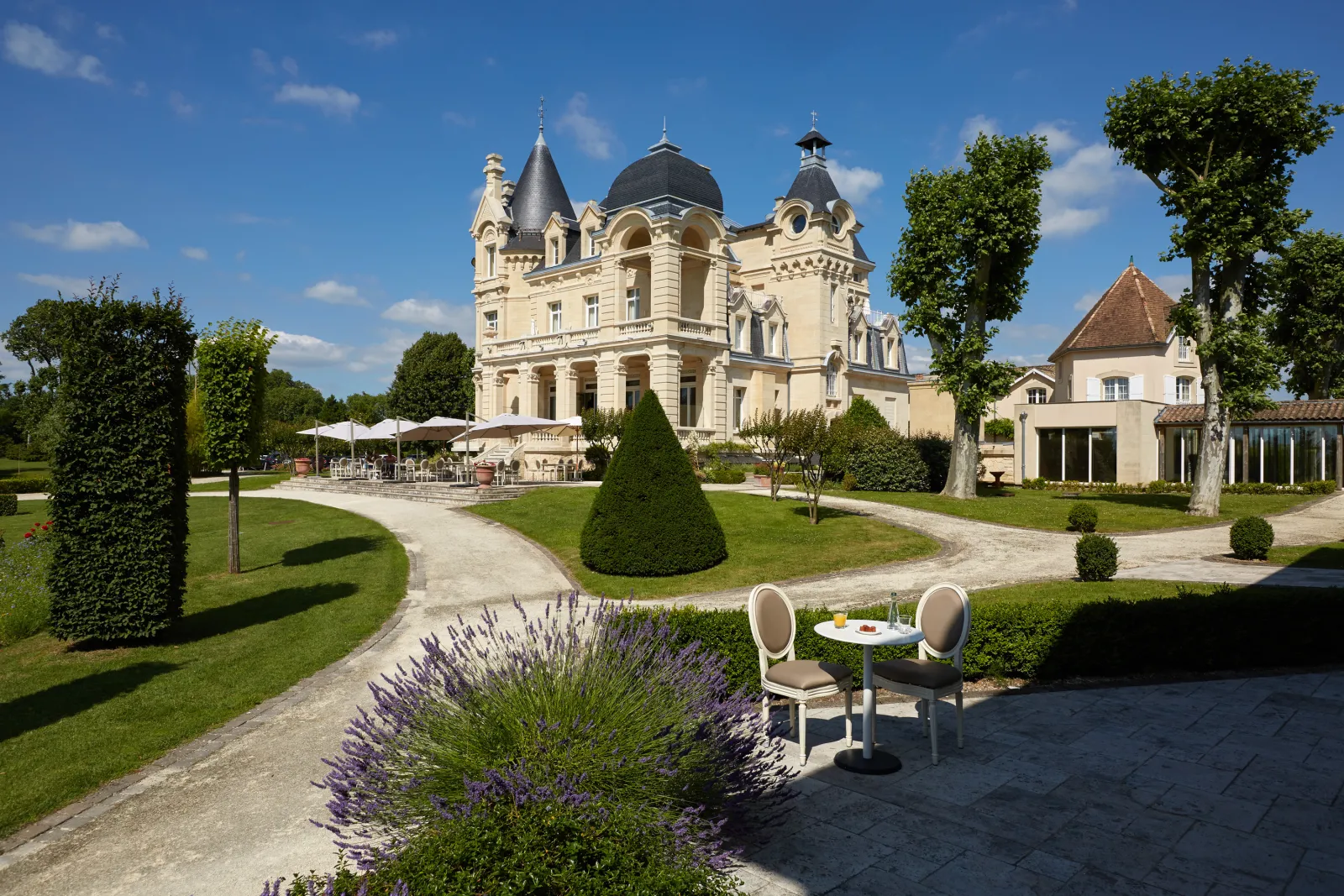 White Belle Époque mansion with black domes surrounded by manicured gardens, lounge chairs in foreground and shaded terrace in background