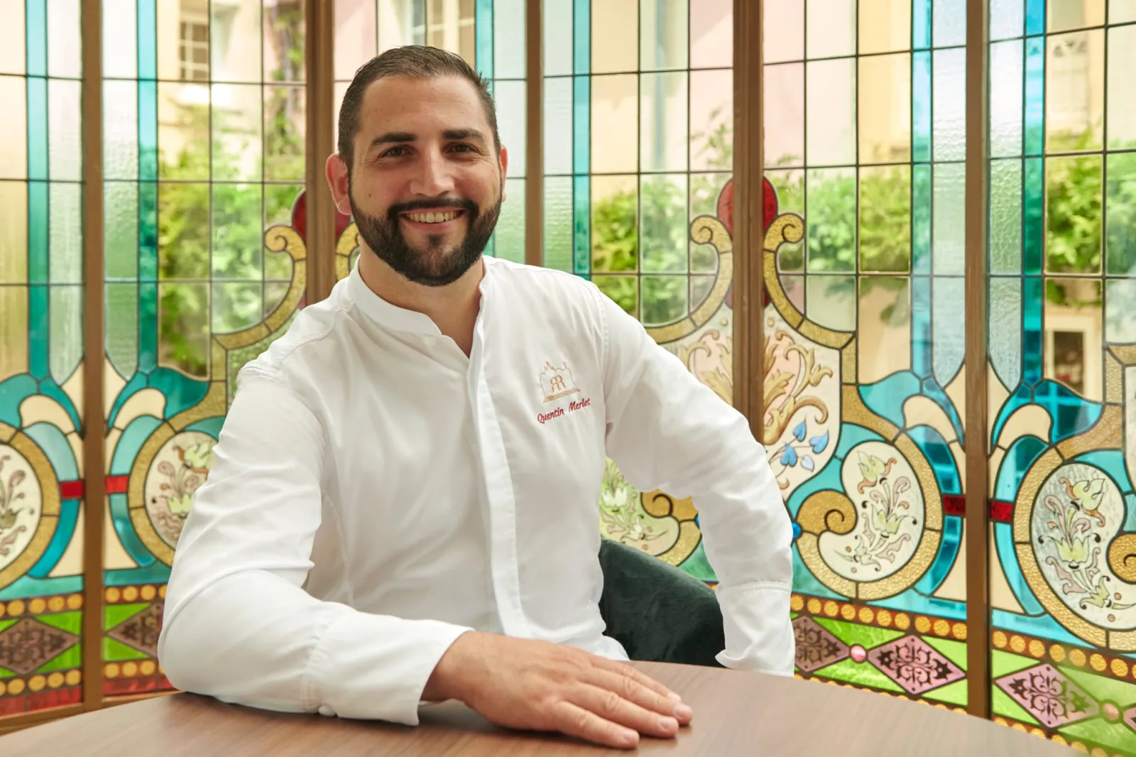 Smiling man in white chef's shirt with restaurant logo, positioned in front of ornate colorful stained glass windows at Grand Barrail restaurant in Bordeaux