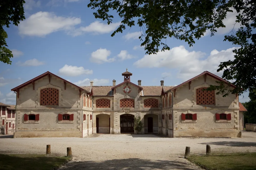 Symmetrical main building of the Horse Museum in Bordeaux, featuring classical architecture with beige stone facade, red tile roof and red wooden doors, set in a paved courtyard.
