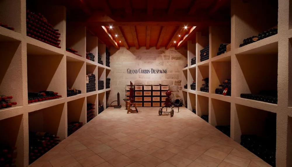 Underground wine cellar of Château Grand Corbin-Despagne in Bordeaux featuring white stone storage racks holding wine bottles, modern lighting and antique furniture in the center
