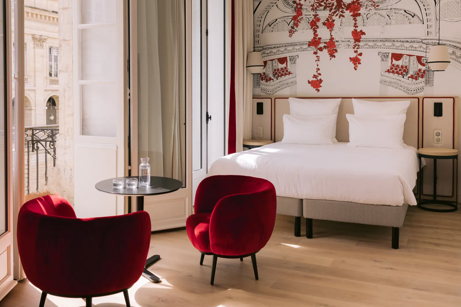 Elegant bedroom at Hôtel Majestic in Bordeaux featuring a white bed, two burgundy velvet armchairs, a balcony overlooking classical Bordeaux architecture, and red floral wall decor.