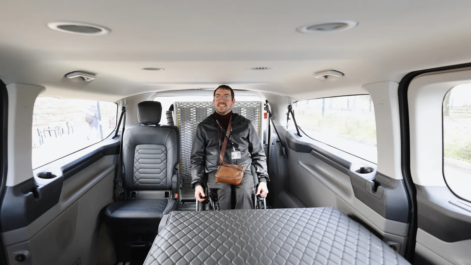 Interior of a transport vehicle with a smiling man wearing a black jacket and brown bag, seated in a spacious cabin equipped with gray seats and a folding bed.