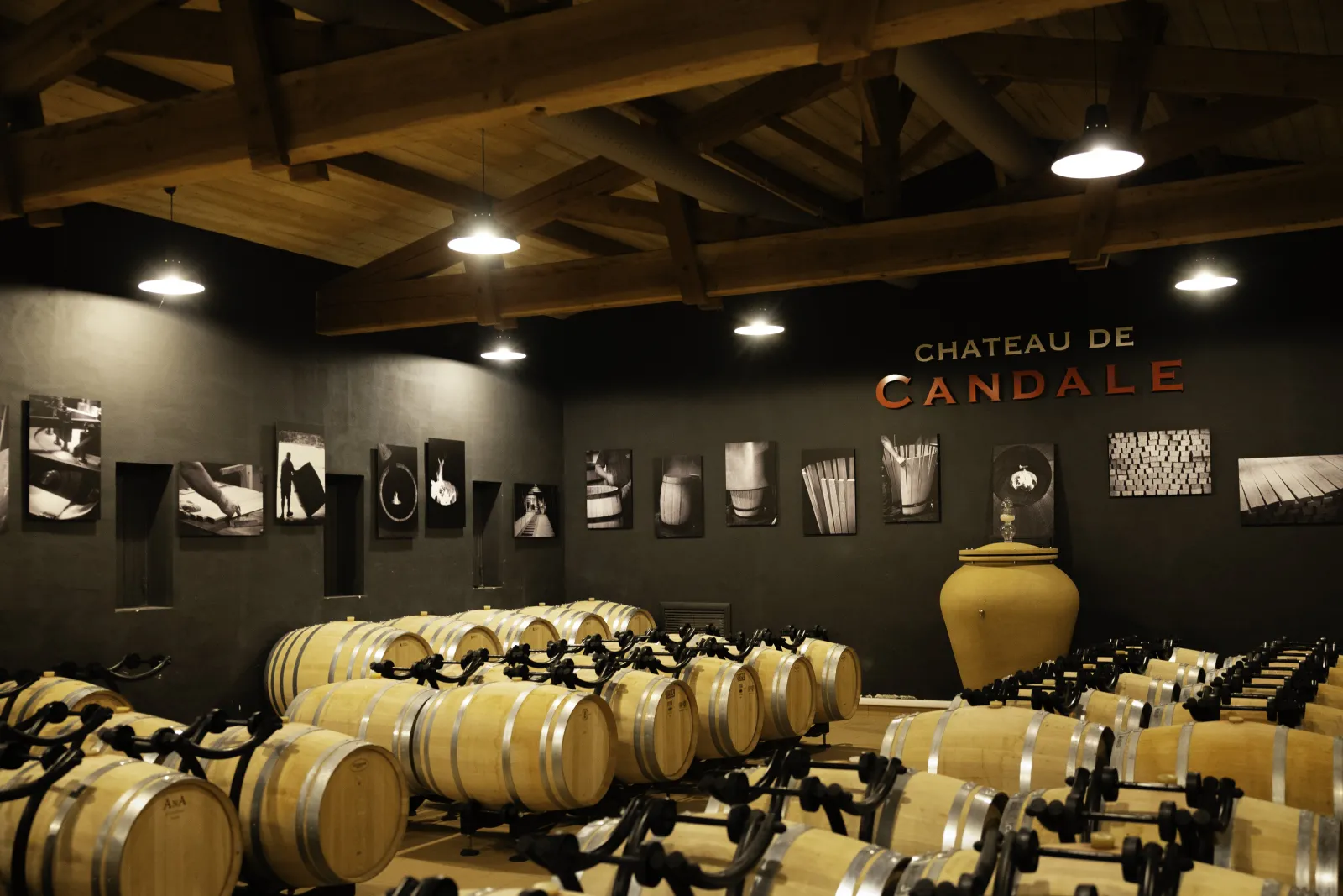 Wine cellar of Château de Candale in Bordeaux featuring wooden barrels in rows, black and white photographic wall display, and a large golden yellow decorative urn.