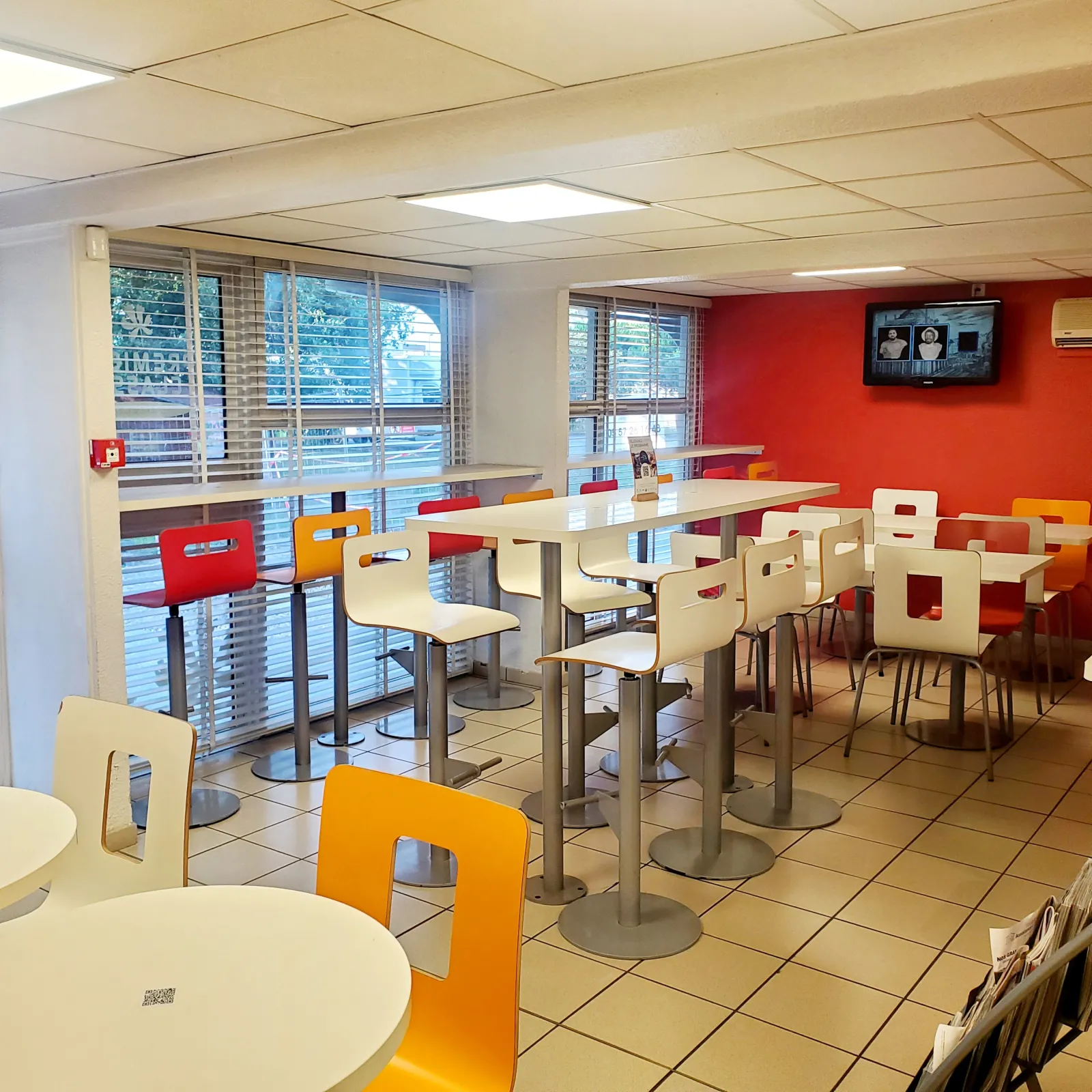 Modern breakfast room at Première Classe Bordeaux South hotel featuring high tables, red and orange chairs, large windows with exterior view, red walls and bright ceiling lighting.