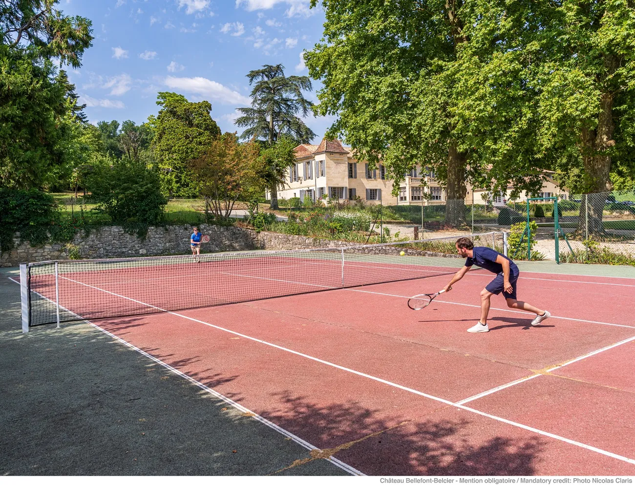A tennis player in action on a red clay court at Château Bellefont Belcier in Bordeaux, with the historic château visible in the background surrounded by trees.