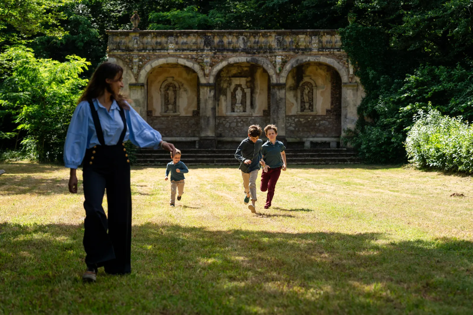 A woman in a blue blouse watches three children running in a park in front of a historic stone building with three arches and sculptured niches, surrounded by greenery at Château de Malle in Bordeaux.