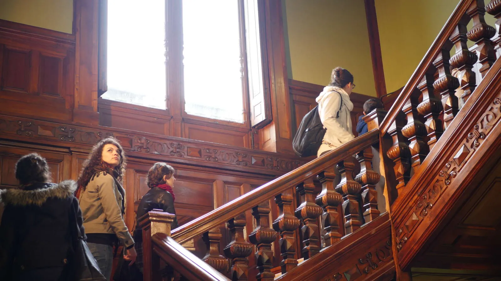 Visitors ascending an ornately carved wooden staircase in a historic building with yellow walls and tall windows, at the National Health Insurance Museum in Bordeaux.