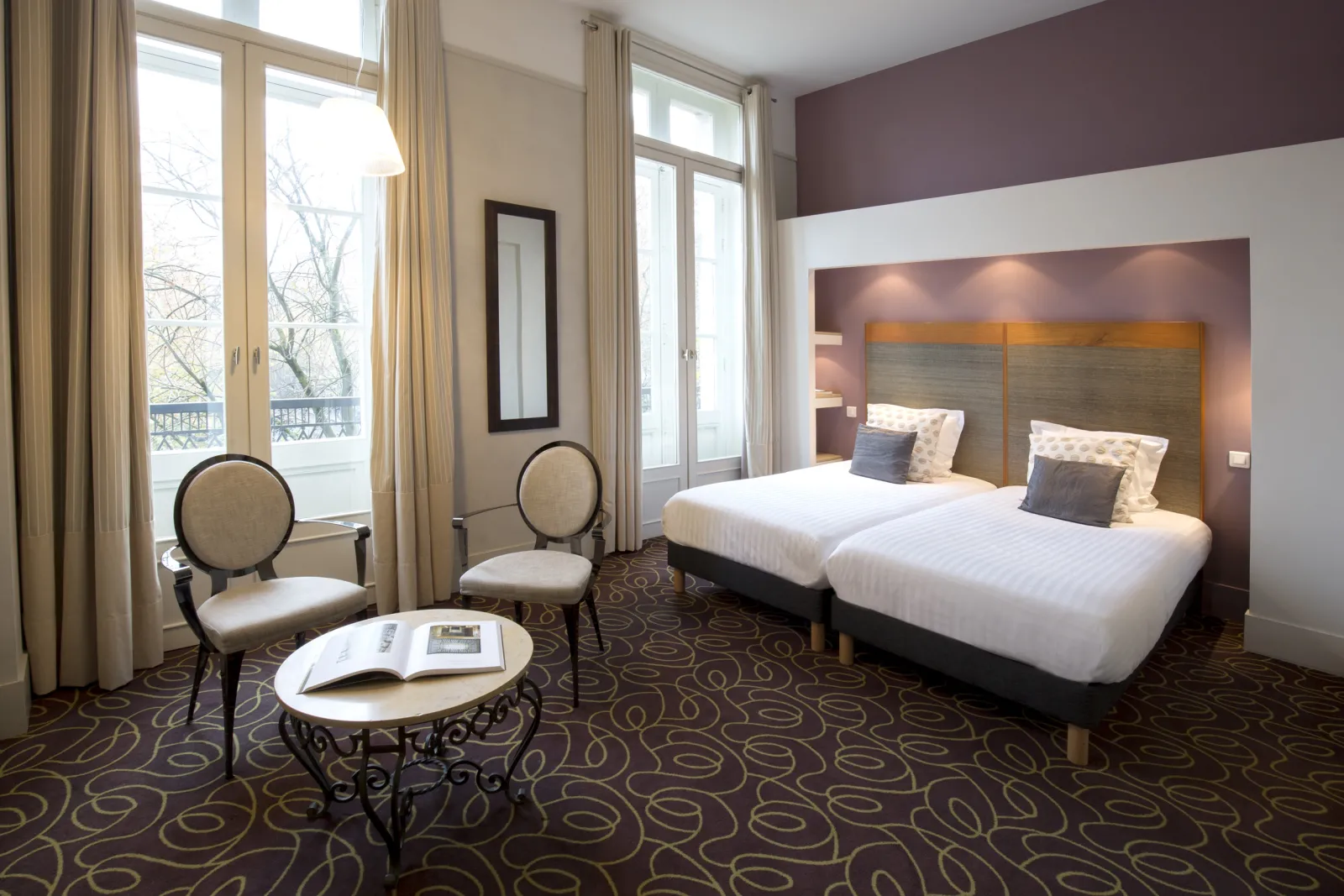 Elegant double bedroom at Hôtel de Normandie in Bordeaux featuring two white beds with wooden headboard, burgundy and white two-tone walls, classic furniture, large windows with balcony view, and patterned carpet.