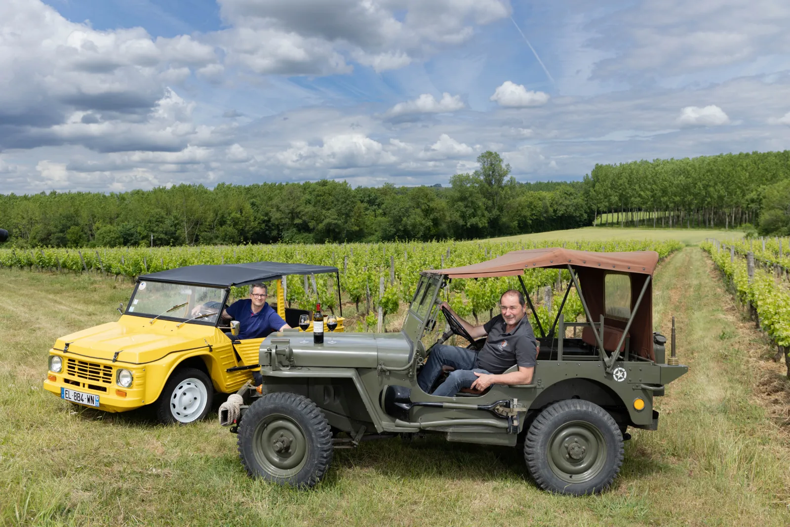 Two men pose with vintage vehicles in Bordeaux vineyards, a green military jeep and a yellow Citroën Méhari, with rows of vines and trees in the background.