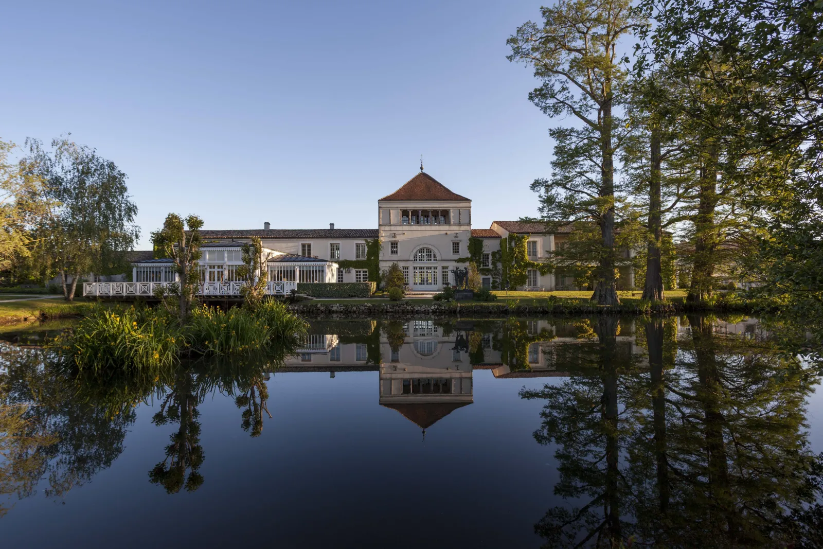 White château with red roofs and central dome perfectly reflected in a calm pond, surrounded by trees and lush vegetation at Les Sources de Caudalie near Bordeaux.