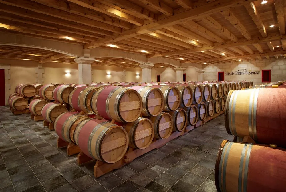 Wine aging cellar at Château Grand Corbin-Despagne in Bordeaux featuring rows of wooden barrels for wine maturation beneath an exposed beam ceiling.