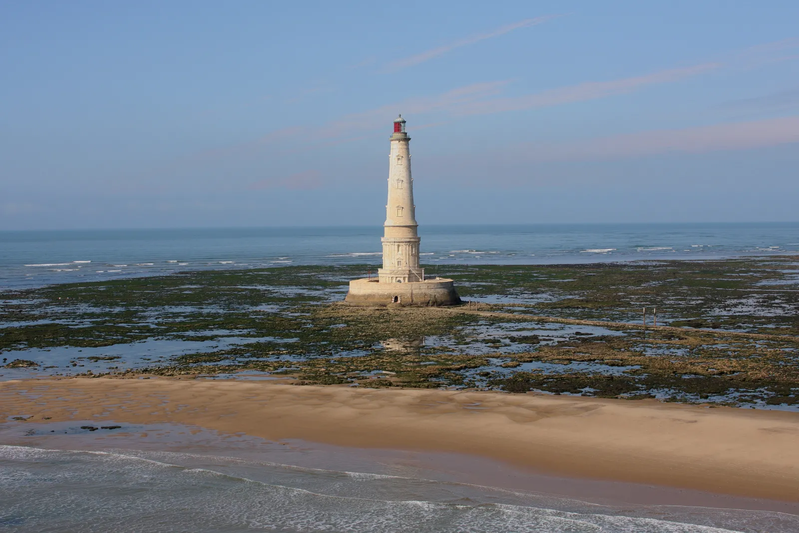 Cordouan lighthouse, a white and beige lighthouse with a red dome, standing on a rocky reef covered with seaweed off the coast of Bordeaux, surrounded by sandy beaches and the Atlantic Ocean.