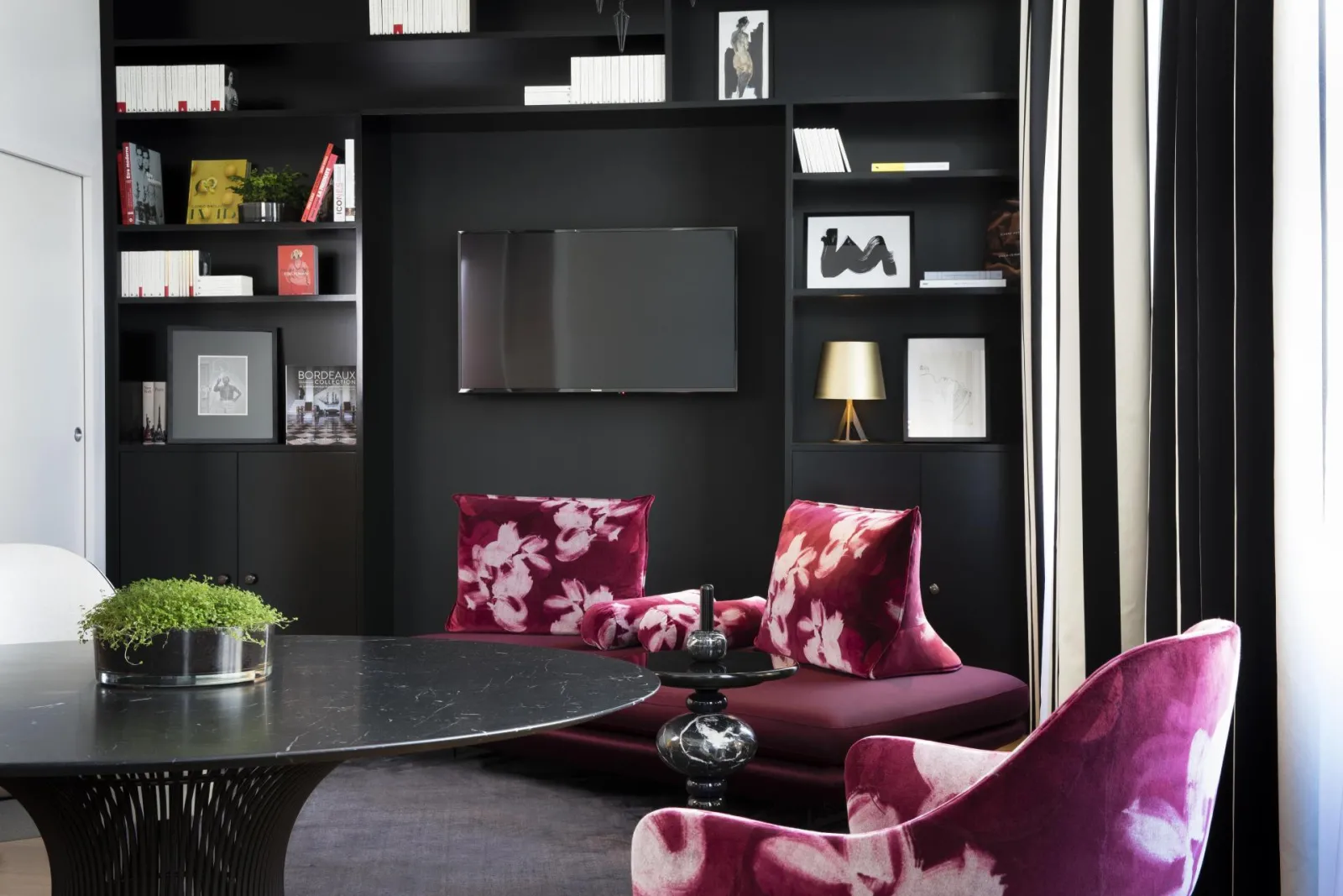 Modern lounge at Hôtel Cardinal Bordeaux featuring black wall with built-in shelving, flat-screen TV, and pink velvet floral armchairs
