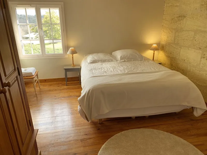 Elegant bedroom at Château La Gontrie in Bordeaux featuring a double bed with white linens, beige stone walls, windows overlooking the garden, gold bedside lamps, and light wood flooring.