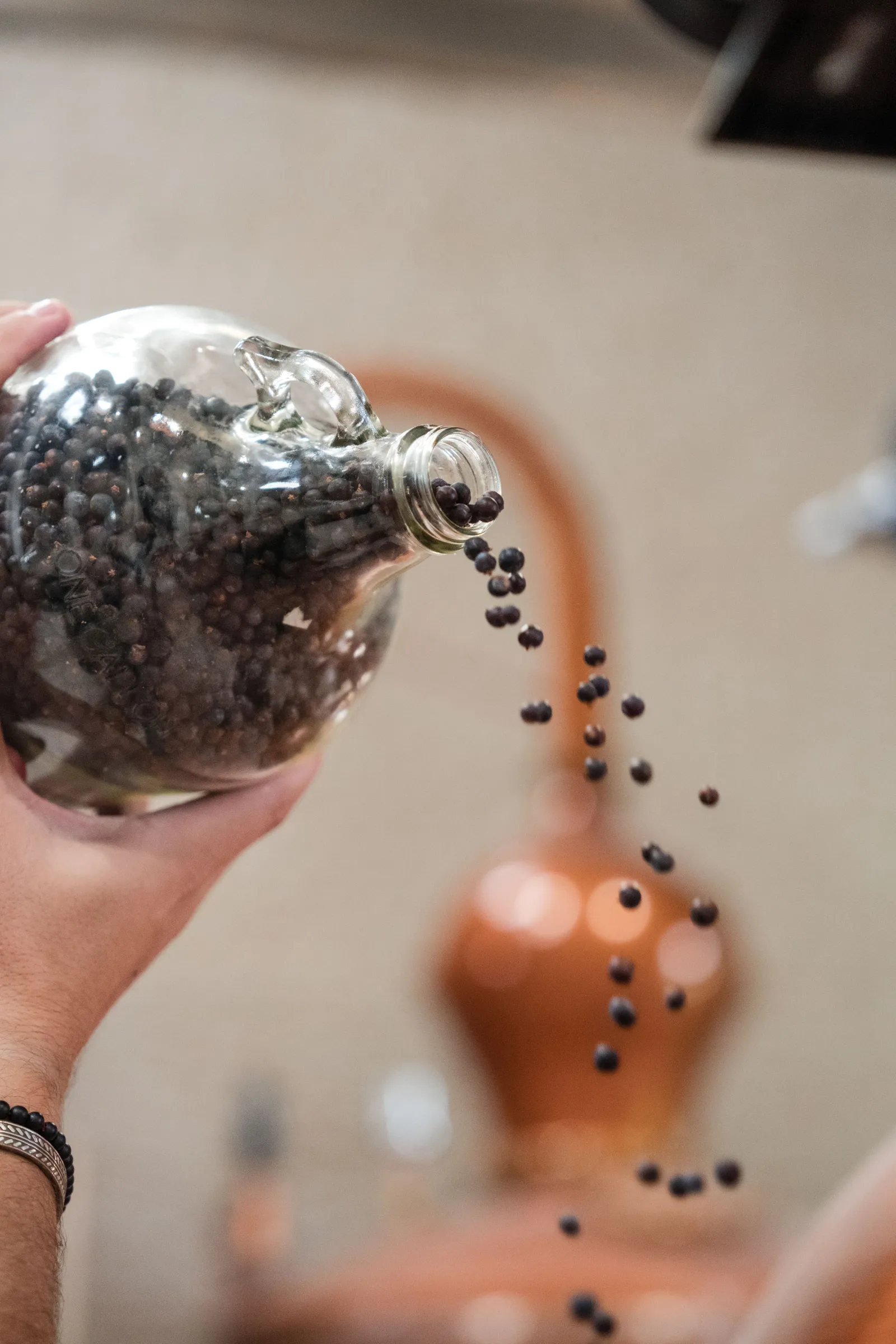 A hand pouring juniper berries from a clear glass bottle into a terracotta ceramic container, demonstrating the traditional craft gin-making process at Citadelle Distillery in Bordeaux.