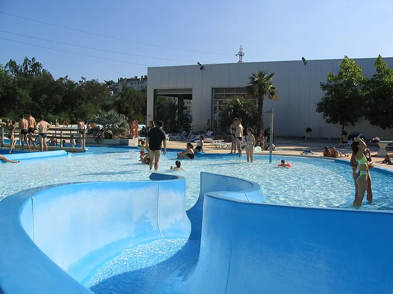 Aerial view of Pessac aquatic stadium in Bordeaux featuring multiple blue swimming pools with visitors swimming, bathing areas, and a white building in the background on a clear day.