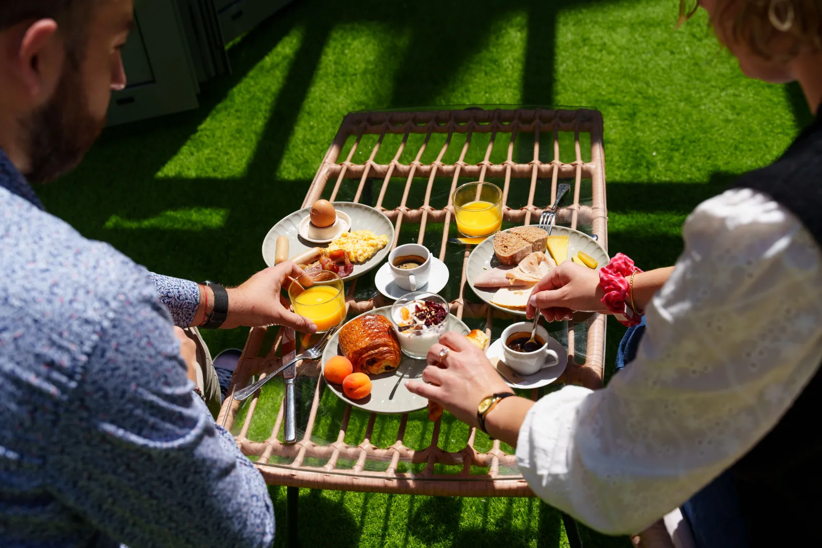 Two people enjoying breakfast outdoors on a wicker table in a sunny garden, with croissants, fresh fruits, orange juice and coffee.