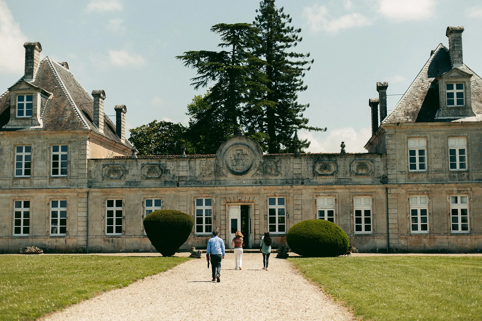 Front facade of Château de Cérons in Bordeaux, a stone building with mansard roofs, visitors walking along the main pathway flanked by trimmed hedges.