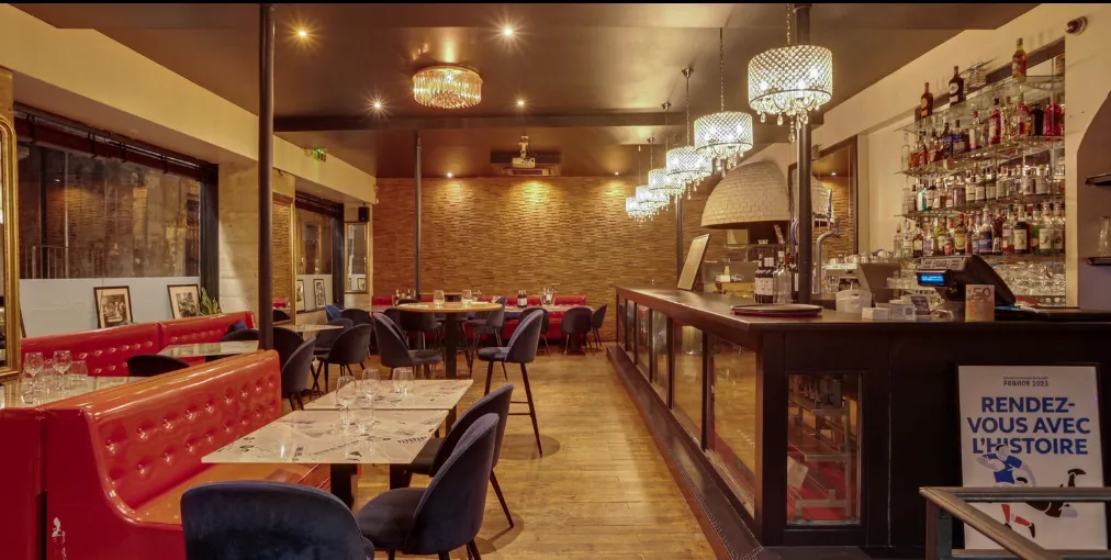 Elegant interior of a Bordeaux restaurant featuring red leather banquettes, dark wood bar, crystal chandelier, set dining tables, exposed brick walls and warm lighting.
