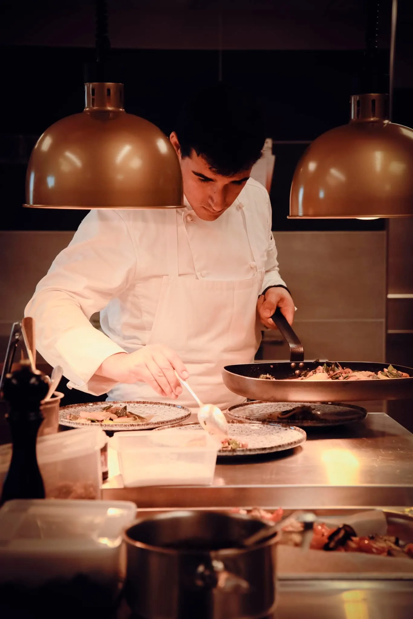 Chef cooking at Magnus table in Bordeaux, preparing a dish under copper kitchen lamps in a modern kitchen.