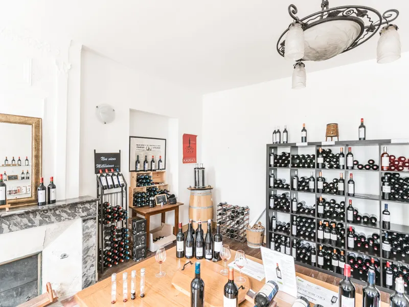 Interior of a wine cellar with tasting counter, metal shelves filled with wine bottles, wooden barrels, tasting glasses and promotional displays in a clean white room.