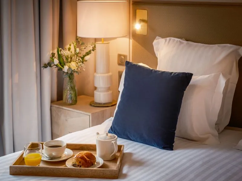 Hotel bedroom with a breakfast tray on the bed featuring coffee, orange juice and a croissant, with bedside lamp, fresh flowers and elegant decor