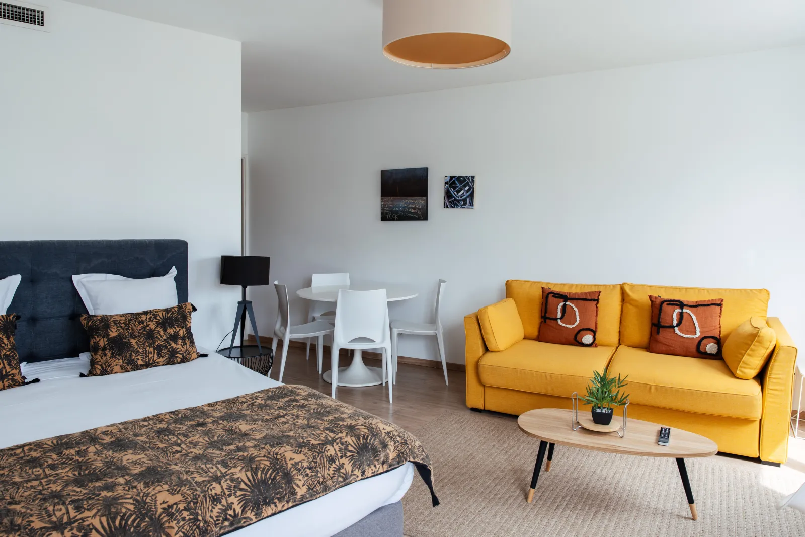 Spacious studio apartment with double bed, yellow sofa, white dining area and golden circular ceiling light at Ténéo Apparthôtel