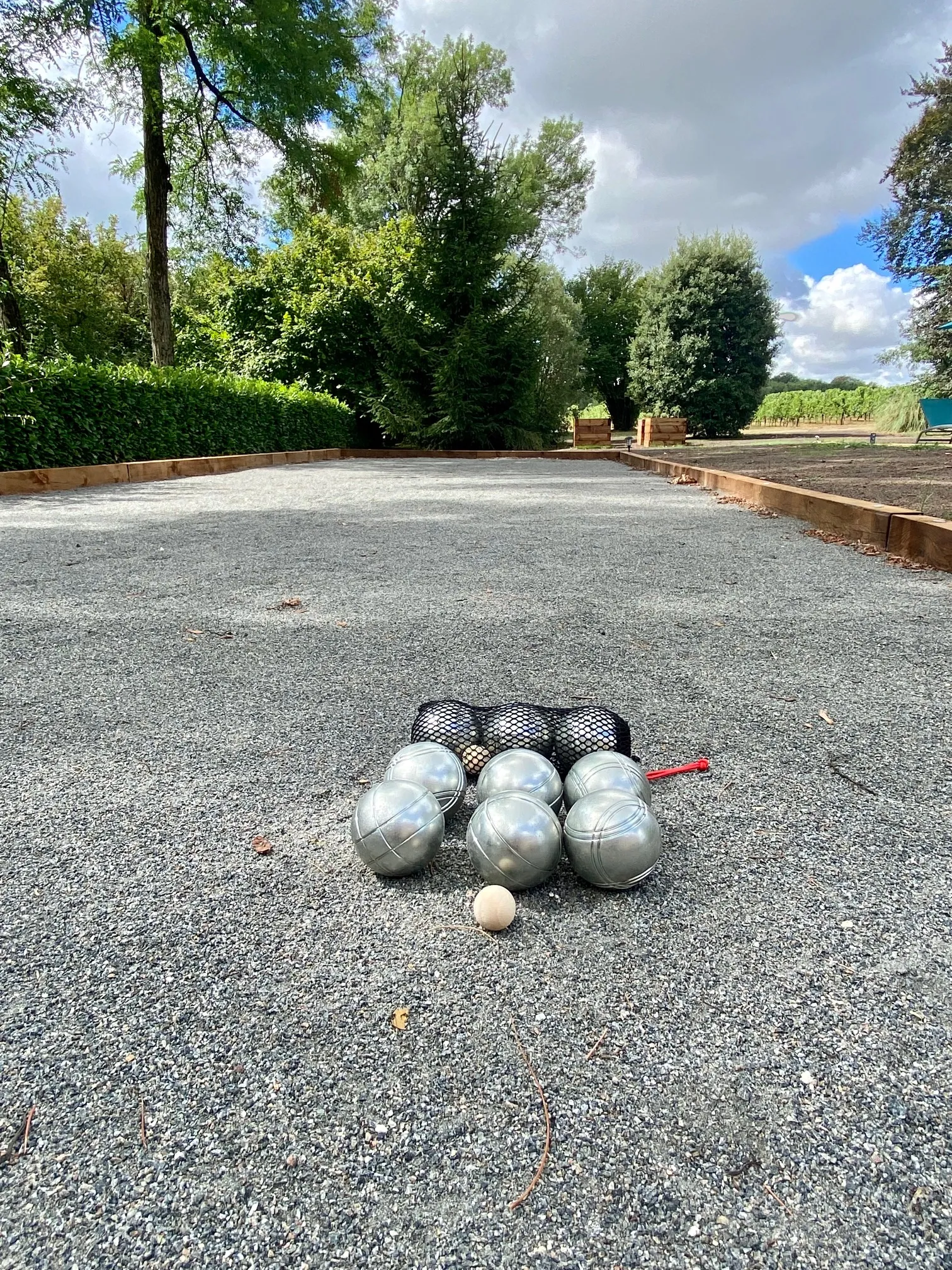 Petanque court with six metal balls in a black mesh bag and a small white target ball on a graveled lane, surrounded by green trees and hedges