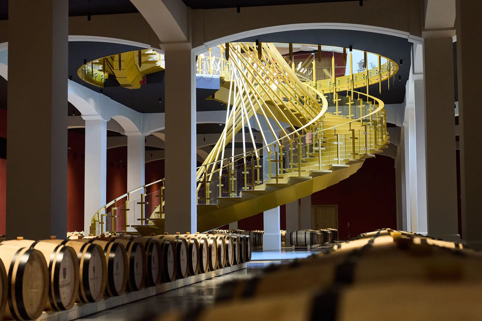Interior of Château Plain Point in Bordeaux featuring a modern yellow spiral staircase, wine barrels in the foreground, and white columns showcasing contemporary winery architecture.