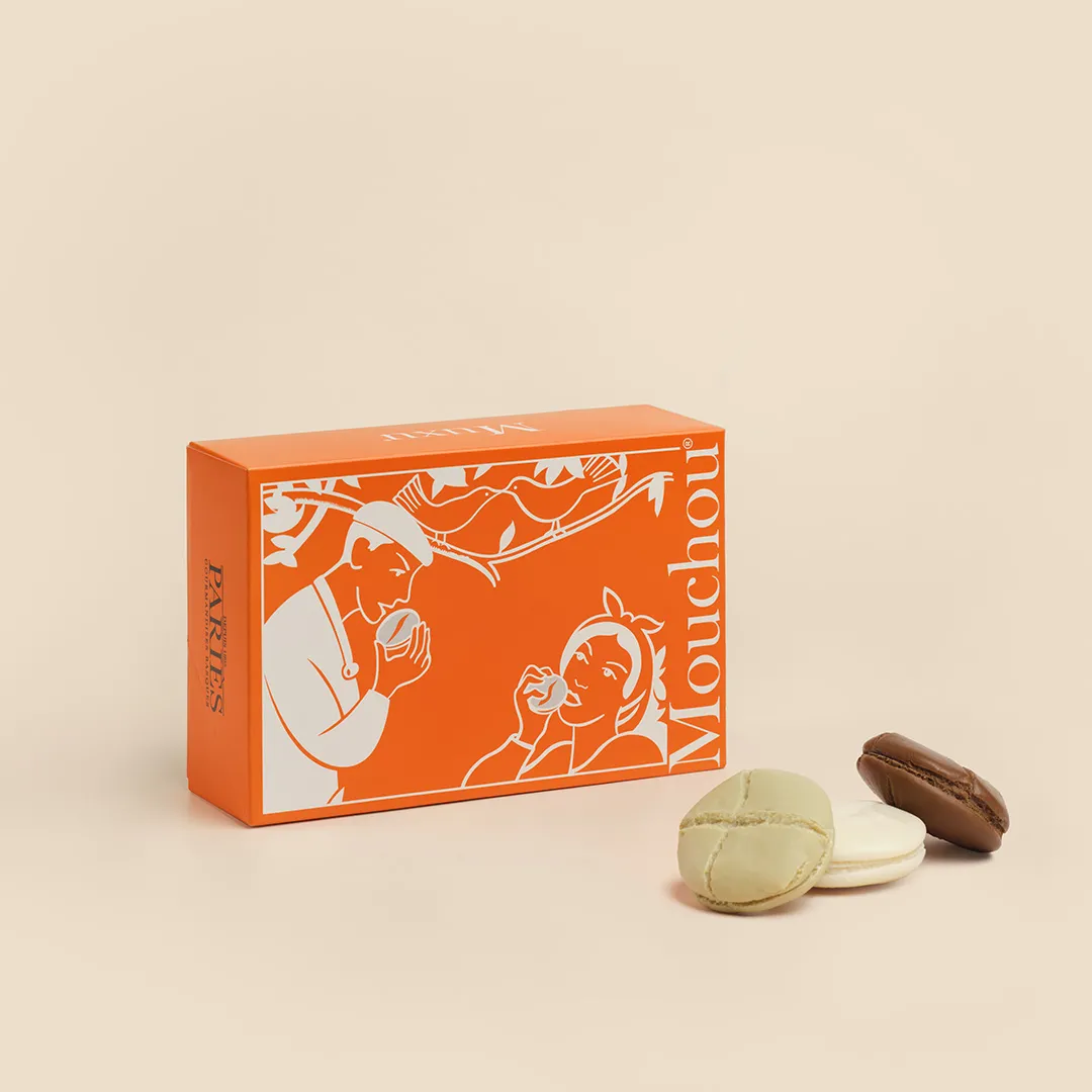 Orange Mouchout macaron box from Bordeaux with white illustrations, displayed with three macarons: two white and one brown, on a beige background.
